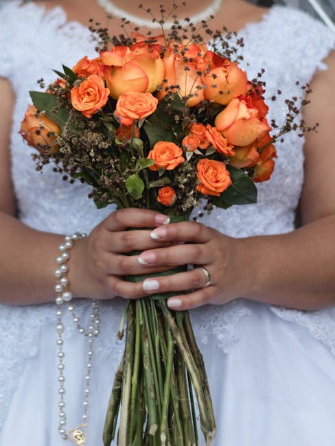 Close-up of a bride holding a bouquet arrangement of peach and orange roses, complemented by small sprigs of baby's breath and dark green foliage. The bouquet features a mix of fully bloomed roses and