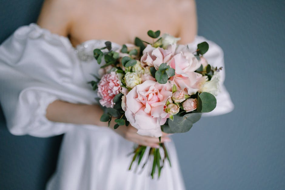 A person dressed in a white off-the-shoulder dress is holding a large bouquet of fresh wedding flowers in front of a dark grey wall. The bouquet features soft pink roses, pale pink and white hydrangea