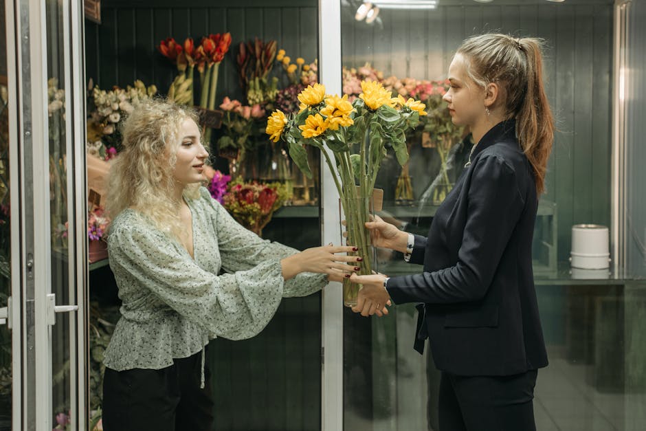 A florist with curly blonde hair, wearing a light-colored, long-sleeved blouse with a ruffled hem, is handing over a bouquet of vibrant yellow flowers, including sunflowers and daisies, wrapped in cle