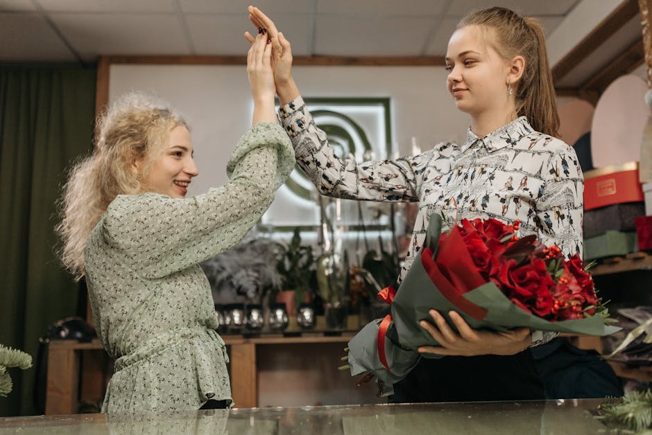 A florist from floristneweltham.co.uk and a young woman are inside a flower shop, exchanging a high-five. The florist holds a large floral bouquet wrapped in dark paper, comprising vibrant red roses,