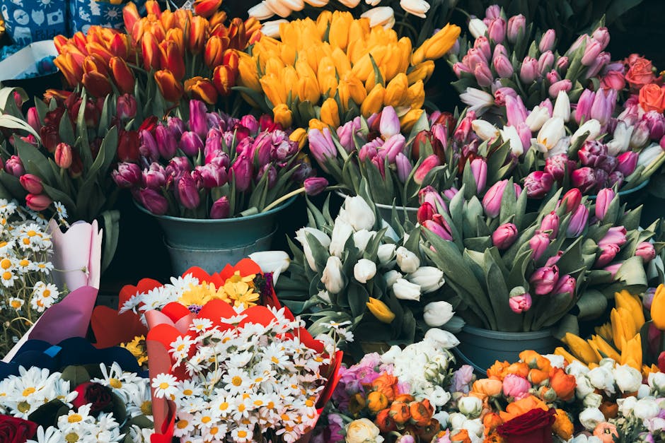 A vibrant display of fresh flowers, including numerous pink, white, and yellow tulips arranged in metal buckets and bouquets. The tulips have partially opened petals, showcasing their delicate texture