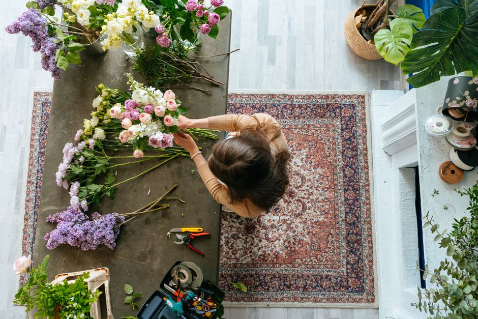 Overhead view of a florist arranging a bouquet of fresh flowers on a dark wooden table inside a well-lit room. The bouquet includes pink roses, white lilies, and purple statice, with some stems and fl