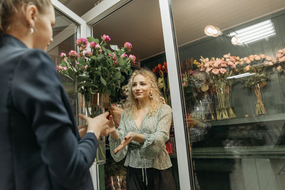 A florist from floristneweltham.co.uk is handing a bouquet of fresh pink roses and greenery to a customer through the open door of a flower shop. The bouquet is wrapped in transparent cellophane and t