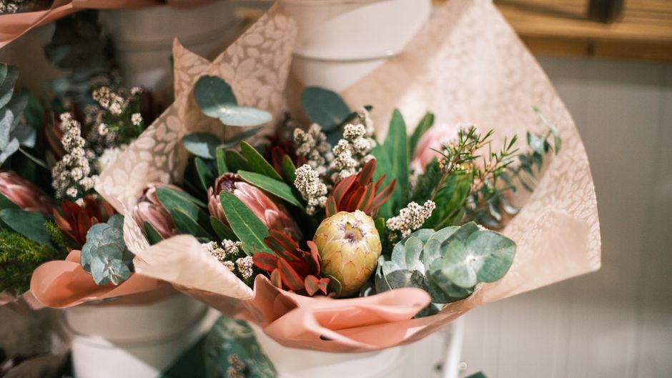 A floral arrangement featuring a mix of fresh flowers and greenery, including pink and red protea, white baby's breath, and eucalyptus leaves, wrapped in soft pink tissue paper with a decorative patte